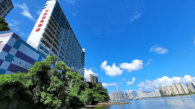 Tall waterfront building with trees, river, and city skyline under a clear blue sky.