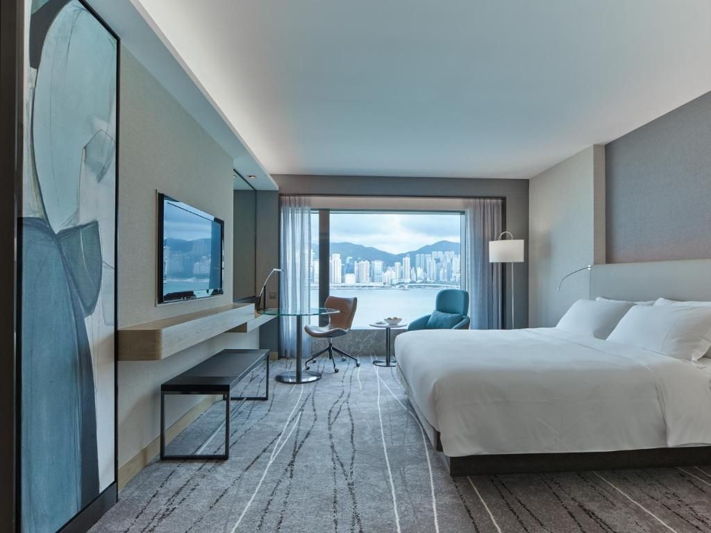 Modern hotel room with king-sized bed, TV, and large window offering city skyline view.