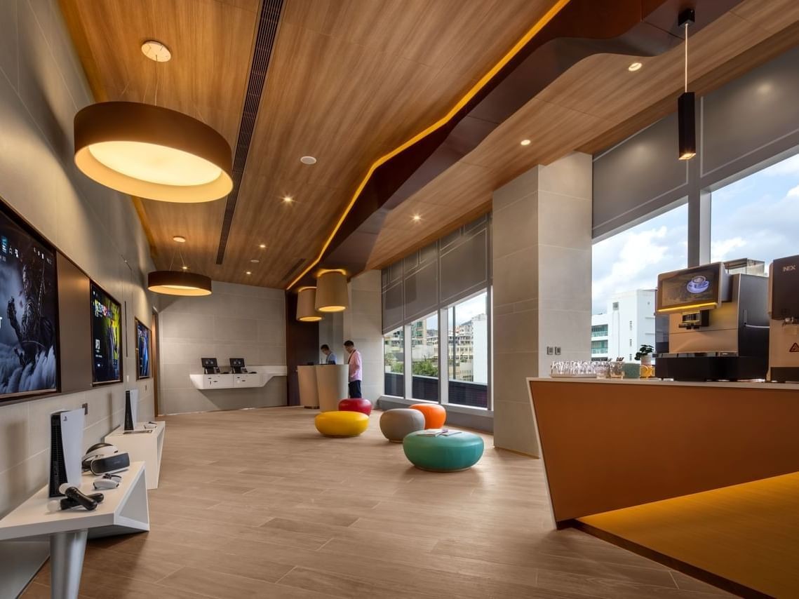 Modern workspace with wooden ceiling, large windows, comfortable seating, gaming setups, and coffee station.