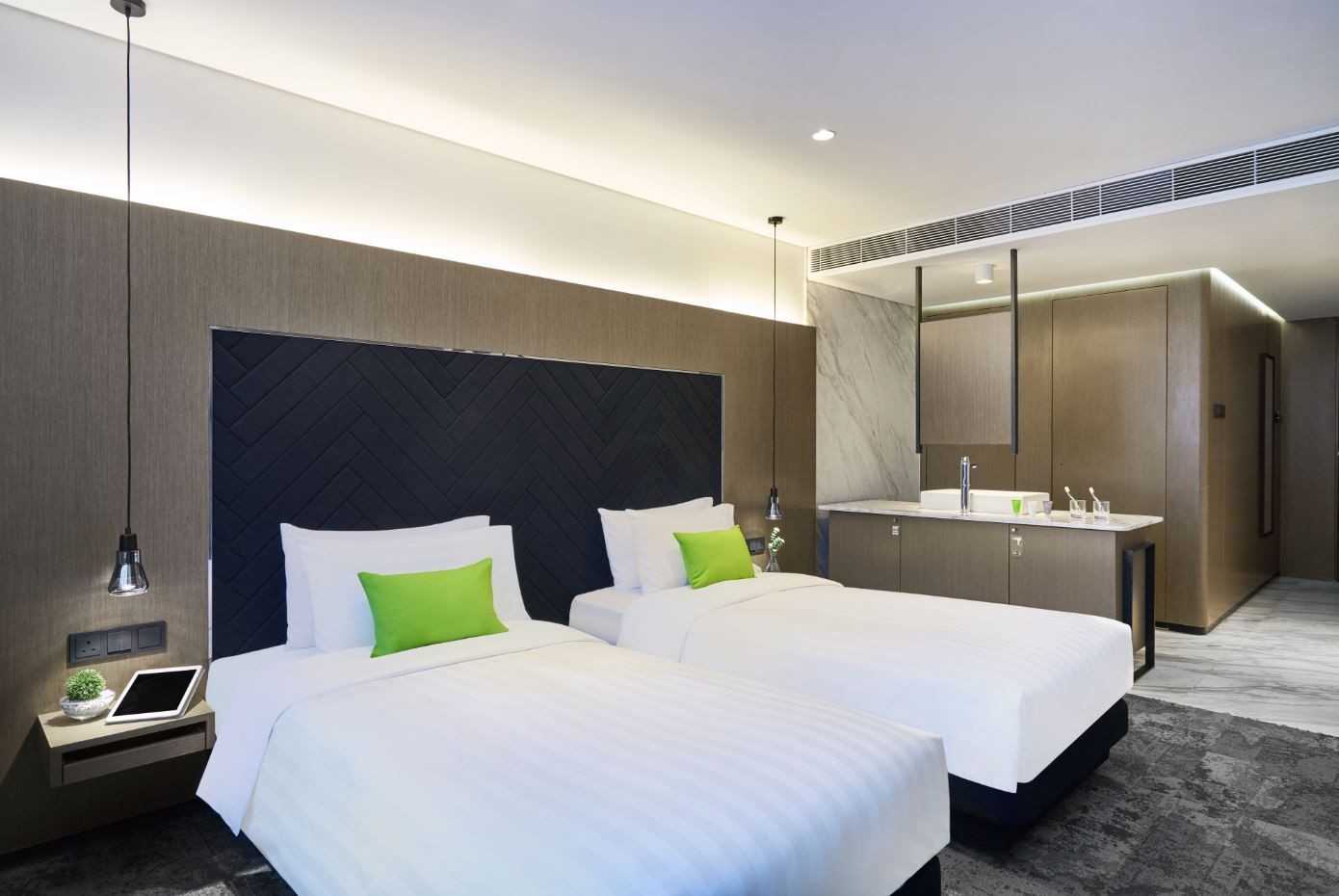 Modern hotel room with two single beds, green pillows, and a kitchenette area.